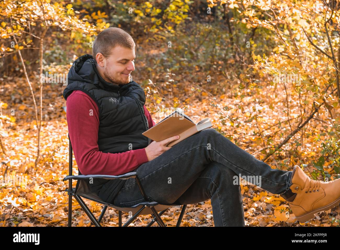 Il giovane è seduto su sedia in un parco autunnale o in una foresta e libro di lettura. Ricreazione mentale in natura. Tranquilla vita di campagna. Escapismo. Disintossicazione digitale. Concetto di lettura del libro in un parco autunnale Foto Stock
