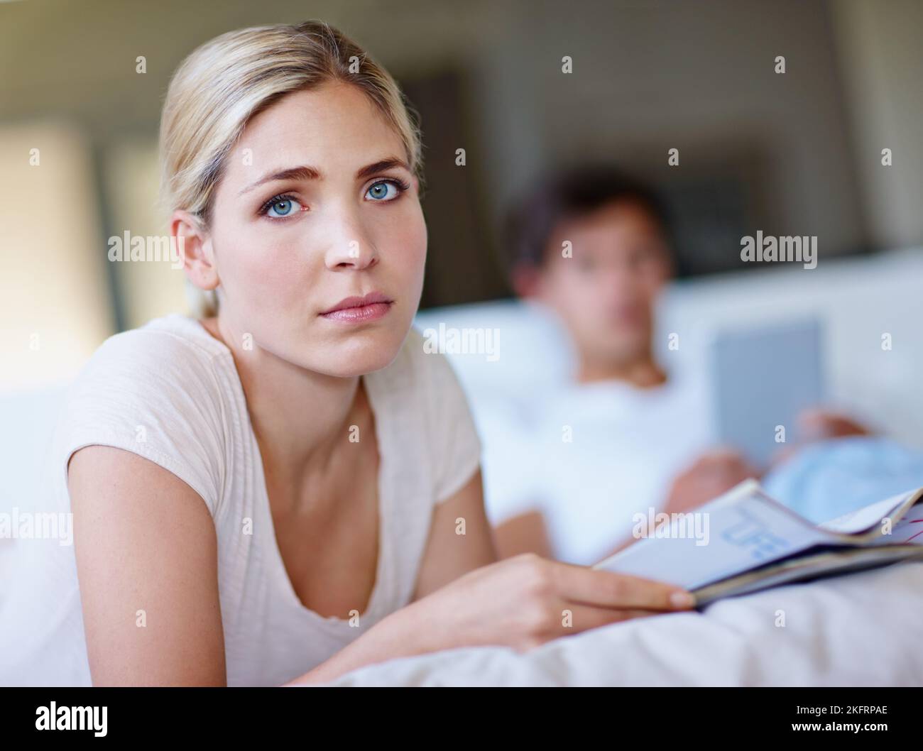 Insieme, ma da sola, una giovane donna triste, sdraiata a letto e leggendo una rivista, mentre suo marito usa un tablet digitale sullo sfondo. Foto Stock
