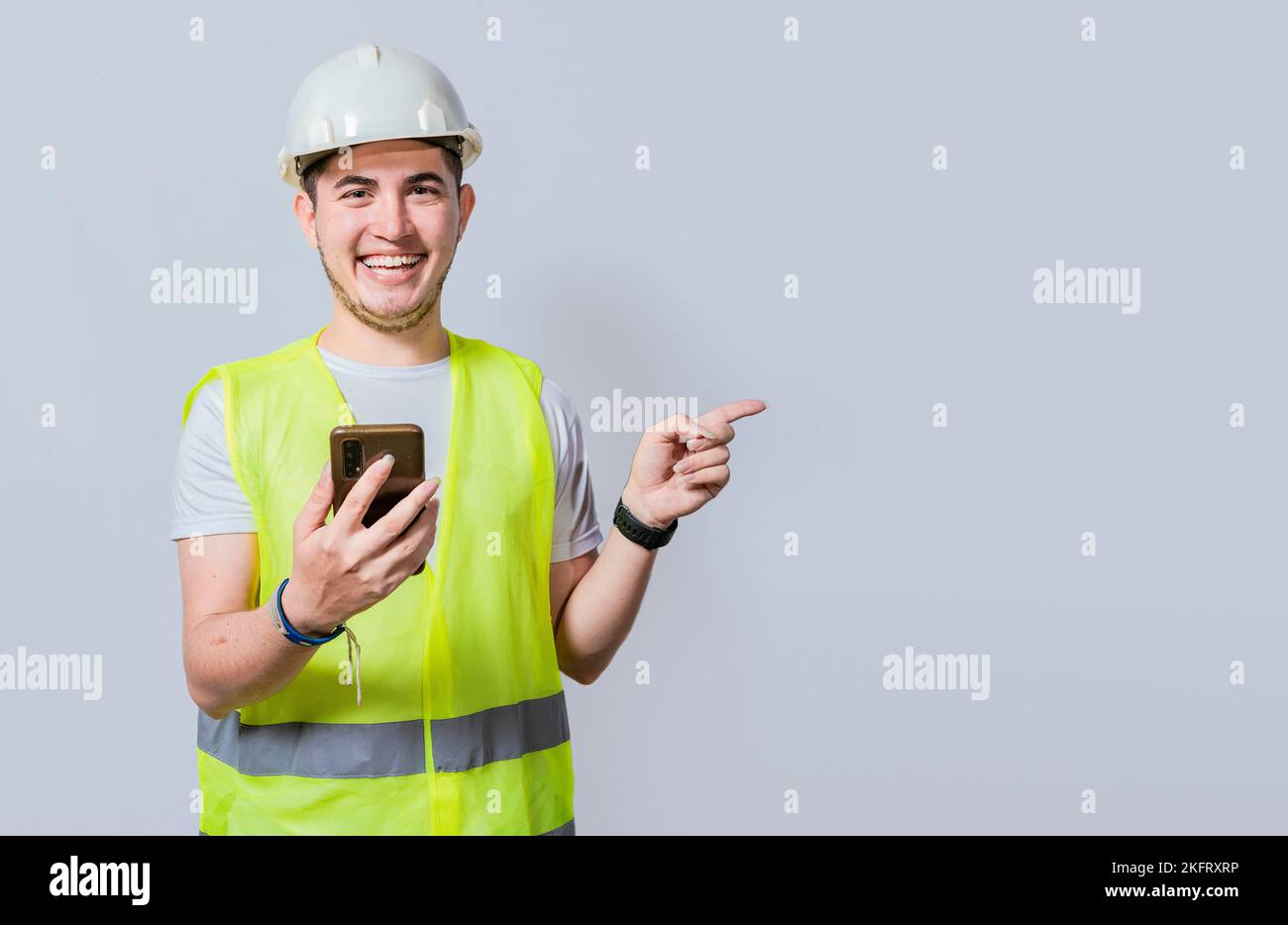 Felice ingegnere civile che tiene il cellulare e punta una promozione. Felice ingegnere che tiene il telefono e che indica al lato isolato. Motore da costruzione giovane Foto Stock