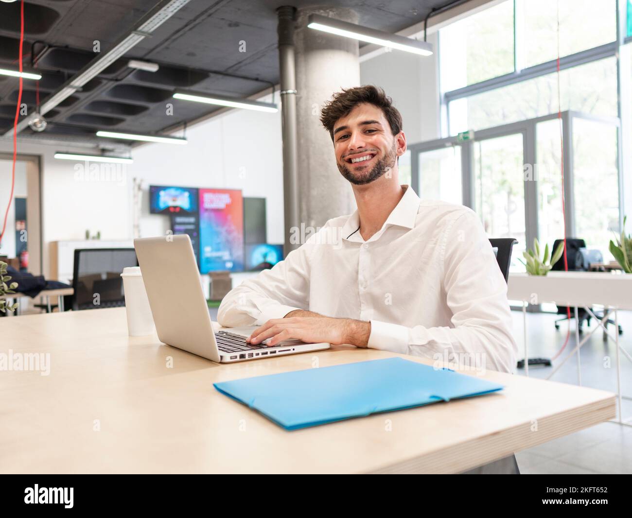 Giovane uomo bearded concentrato in camicia bianca seduto al tavolo e navigando in internet via computer portatile mentre si lavora su un computer portatile in una spaziosa caffetteria ufficio Foto Stock