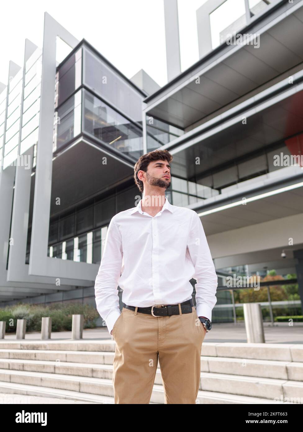 Basso angolo di giovane uomo bearded pensivo in abito casual elegante in piedi vicino a un edificio urbano con le mani in tasca e guardare lontano Foto Stock