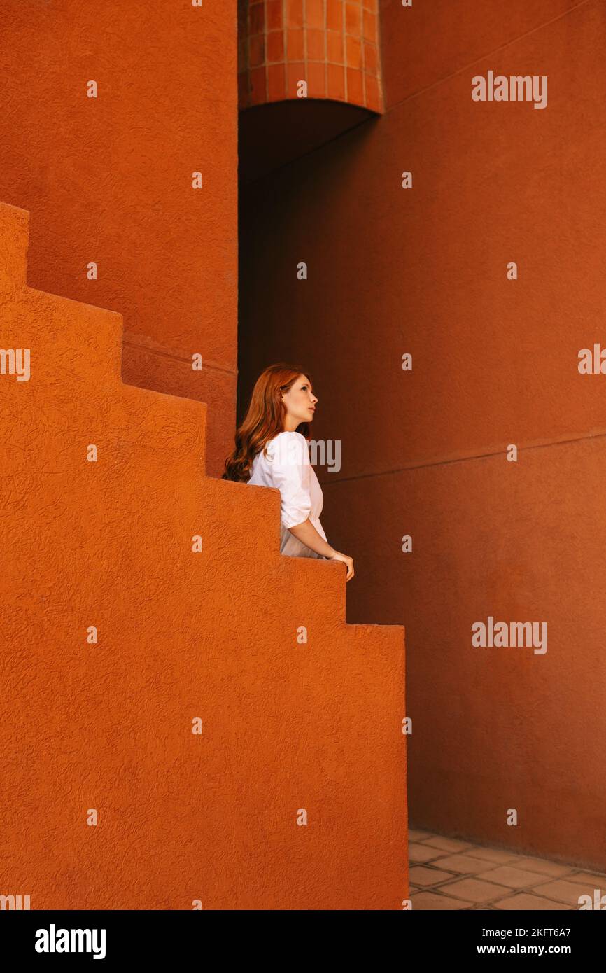 Vista laterale di una giovane femmina rossa in piedi nell'edificio con pareti arancioni vicino alla scala che guarda lontano Foto Stock