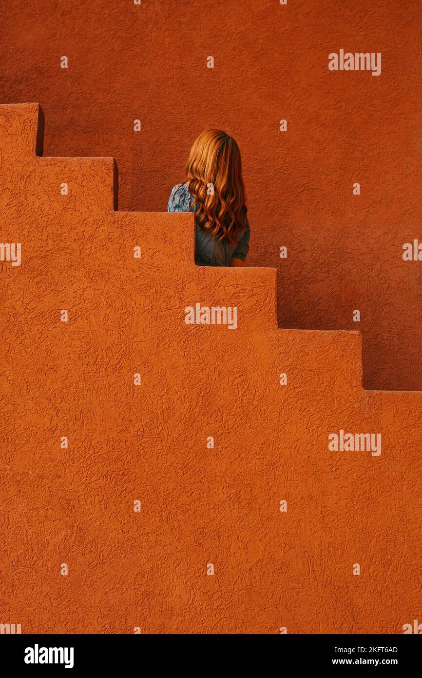Vista posteriore di una giovane donna irriconoscibile rossa in piedi nell'edificio con pareti arancioni vicino alla scala Foto Stock