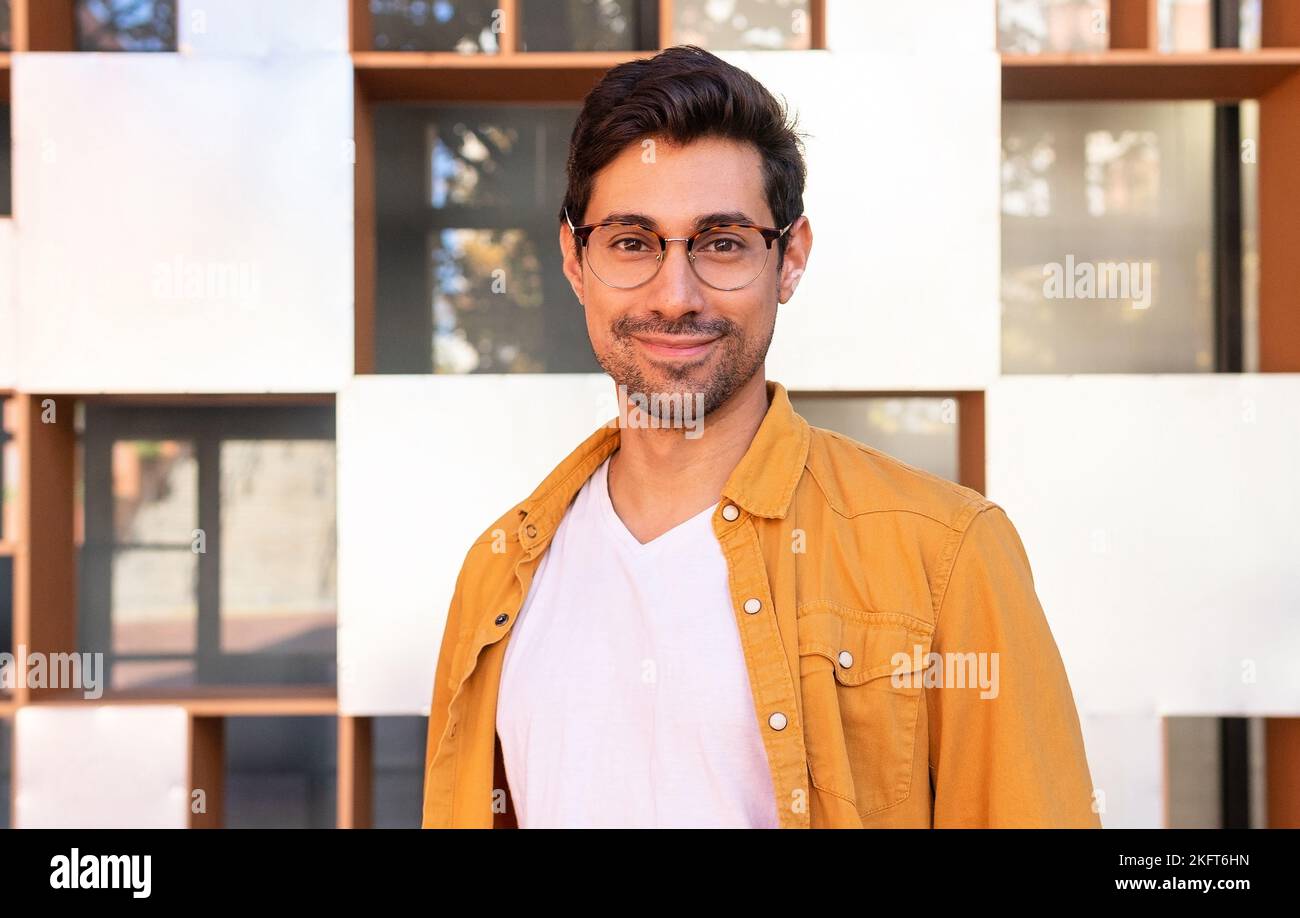 Uomo positivo giovane ispanico sopportato in abiti casual e occhiali in piedi vicino alla parete dell'edificio urbano e guardando la macchina fotografica Foto Stock