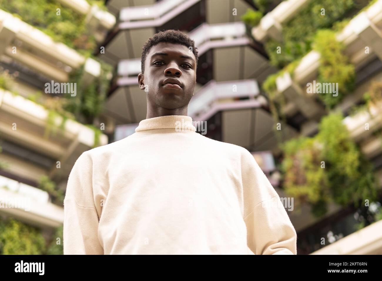Dal basso di un giovane ragazzo afroamericano in abiti leggeri che si erge contro un alto edificio in città e guarda la macchina fotografica Foto Stock