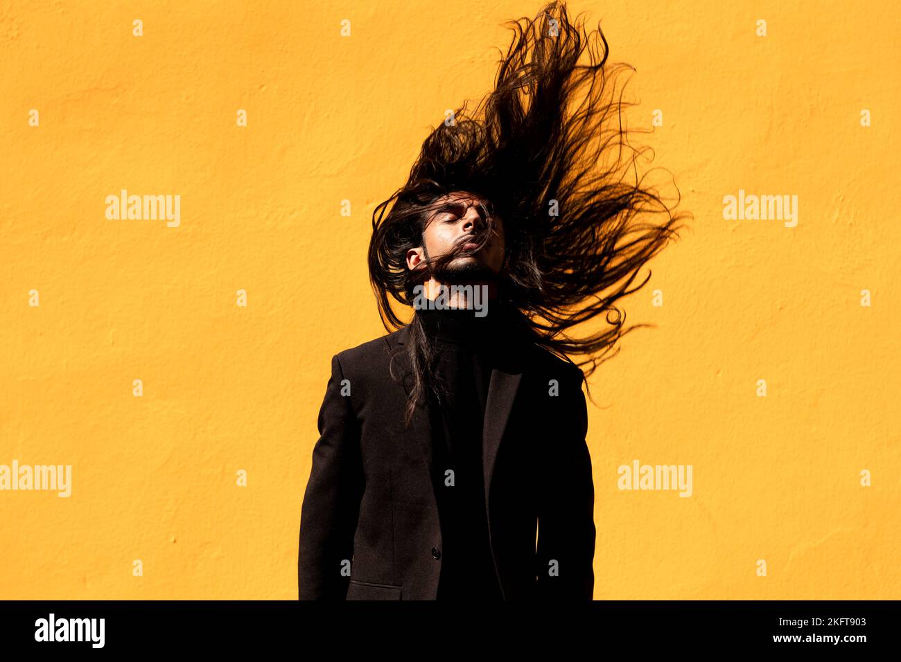 Giovane uomo ispanico in tuta nera che chiude gli occhi e scuotendo lunghi capelli scuri su sfondo giallo Foto Stock