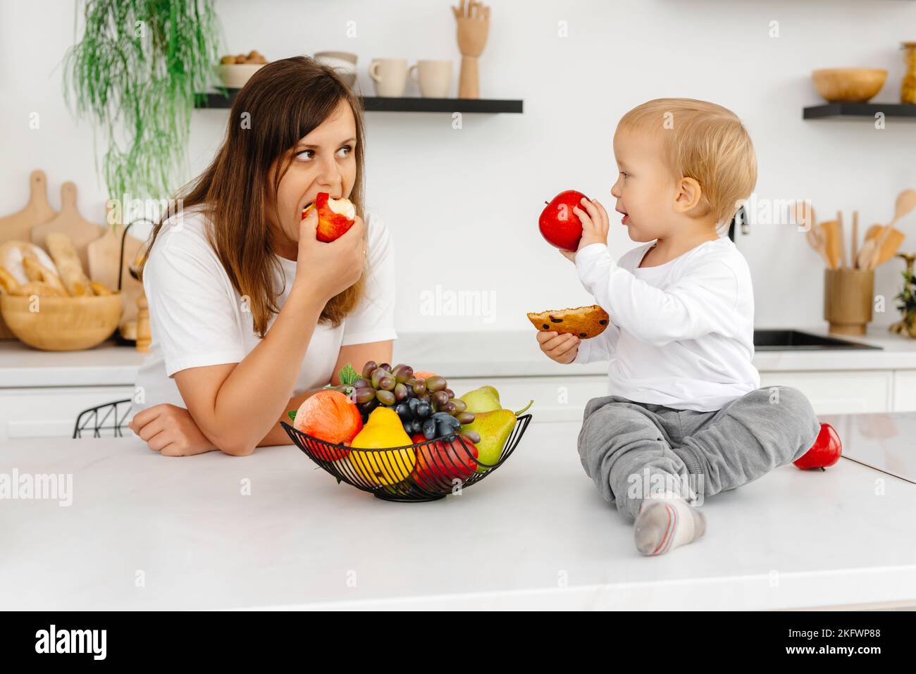 Mamma e suo figlio sono in cucina a mangiare cibo sano. il bambino sta tenendo un biscotto e una mela. Mangiare Reducetarian godendo di cibo a base di piante Foto Stock