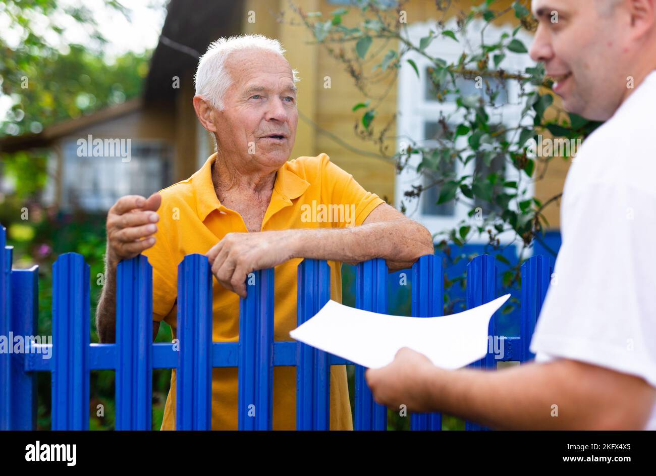 Uomo anziano che comunica con il rappresentante della compagnia di assicurazioni mentre si trova alla recinzione della sua casa di campagna Foto Stock