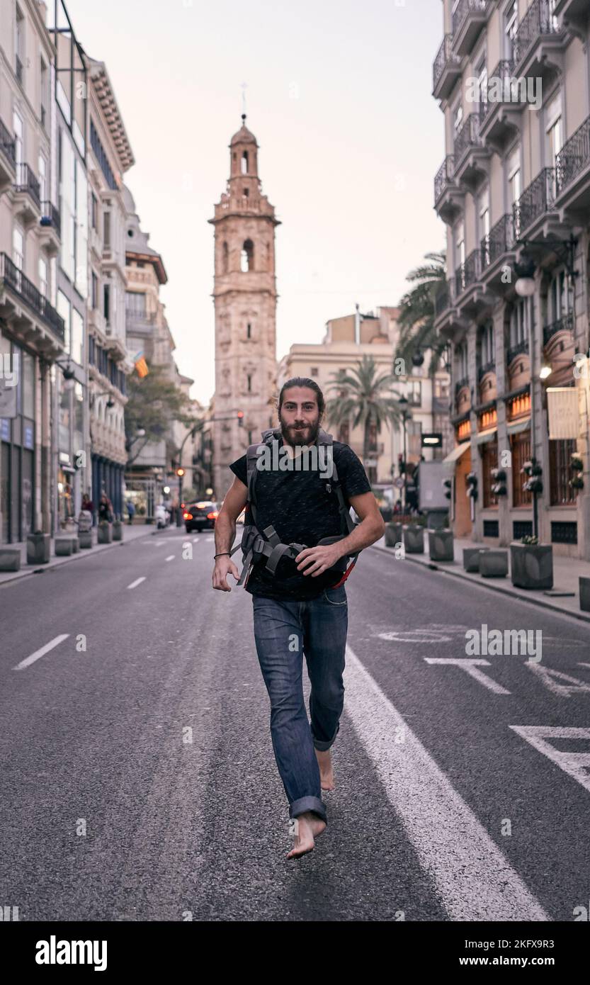 giovane uomo caucasico con grande zaino sulla schiena che corre a piedi nudi verso la fotocamera attraverso una strada cittadina, valencia, spagna Foto Stock