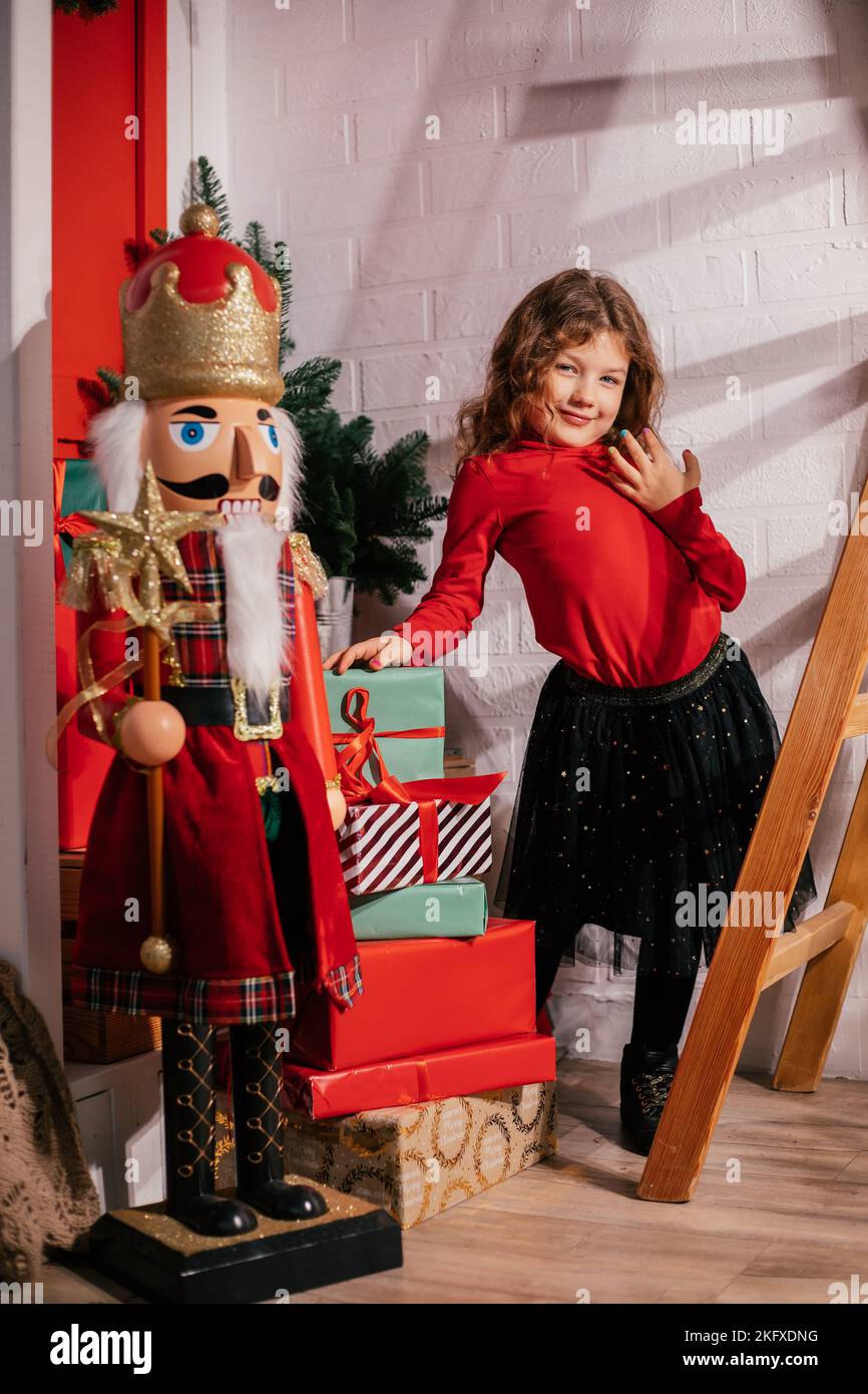 Ragazza verticale sorridente, giocosa, artistica ricci in piedi vicino al grande giocattolo Nutcracker in corona, vicino al presente scatole in negozio di giocattoli Foto Stock