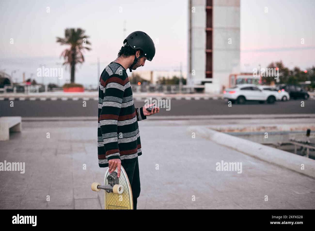 giovane caucasico in pullover a righe e casco protettivo sulla sua testa in piedi guardando il suo smartphone con skateboard in mano Foto Stock