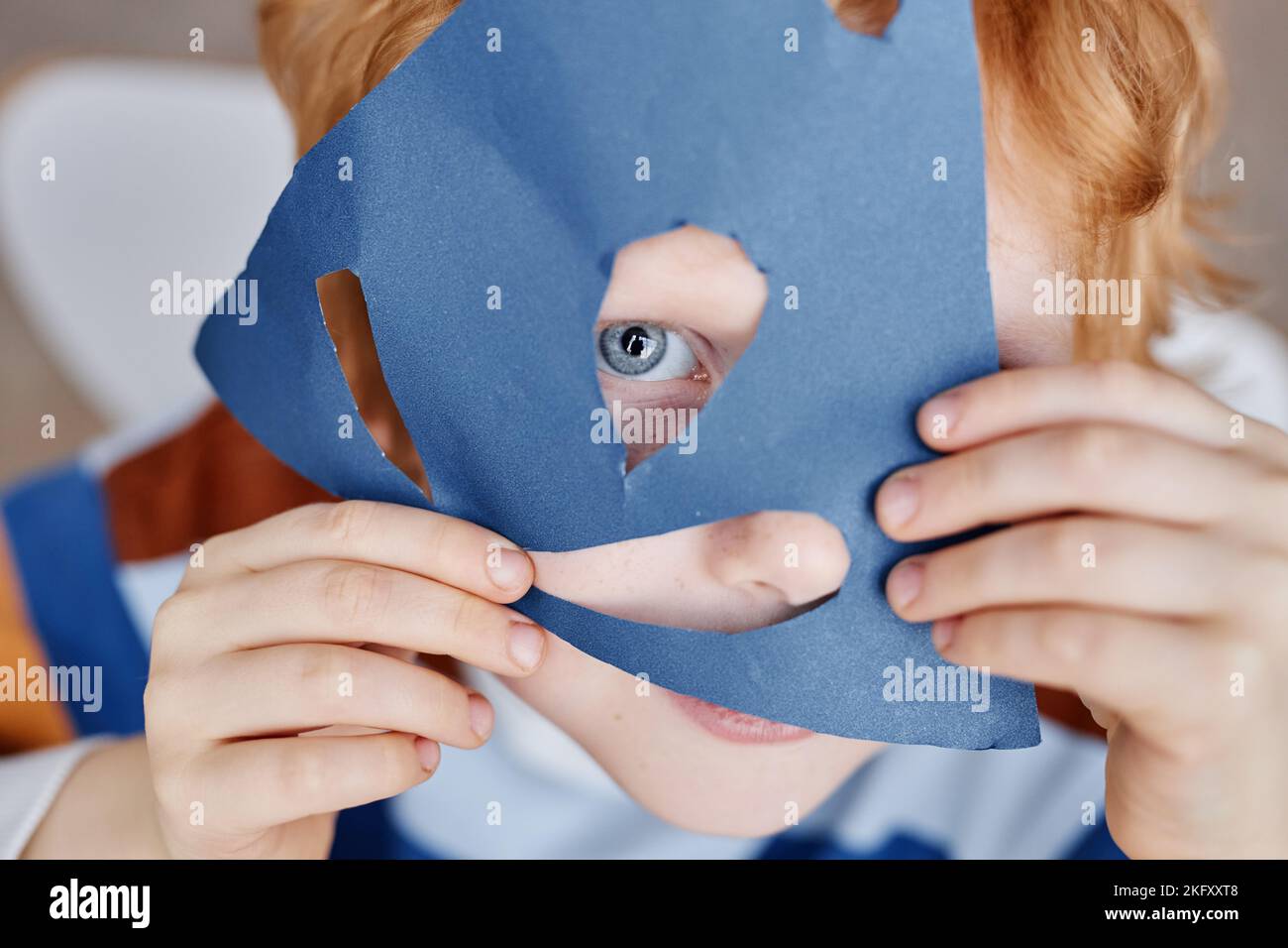 Primo piano di un simpatico ragazzino blueeyed che copre il viso con una maschera di carta blu fatta a mano, mantenendo occhio e naso nei fori tagliati Foto Stock