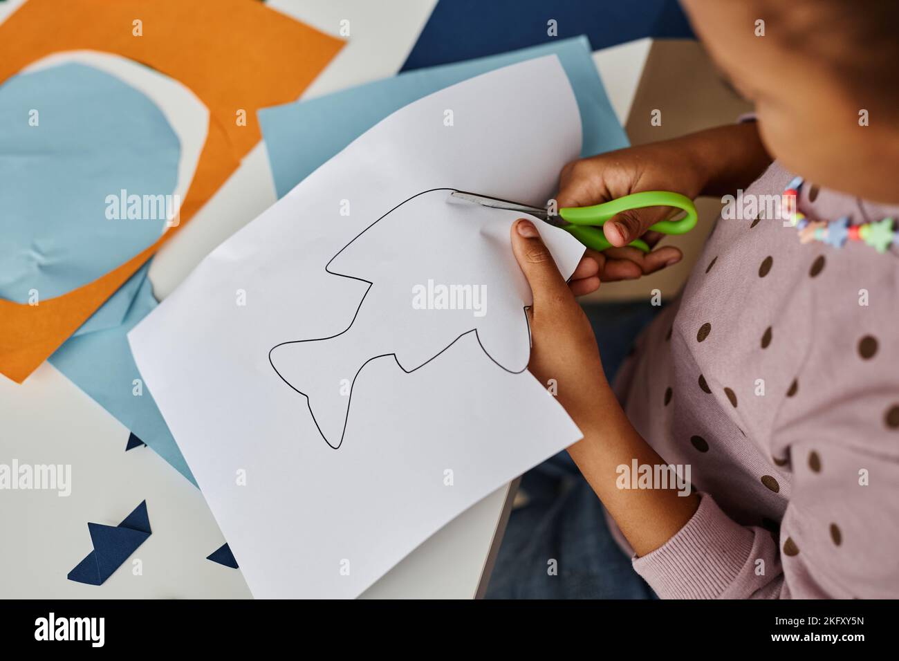Sopra scatto di piccola ragazza afro-americana che taglia il profilo del pesce disegnato su carta bianca mentre genera i giocattoli alla lezione nella scuola materna Foto Stock