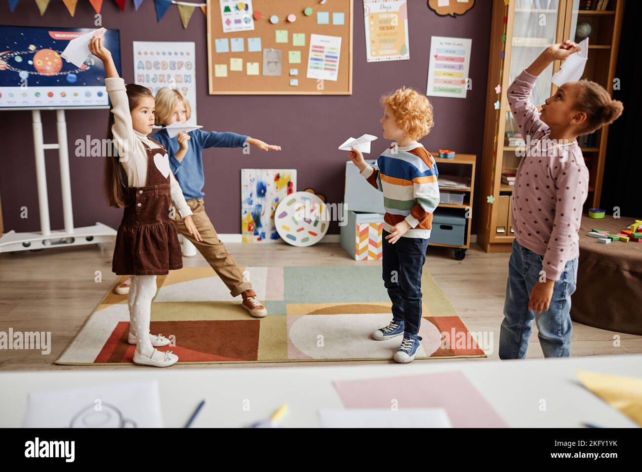 Gruppo di bambini multiculturali che giocano aerei di carta fatti a mano e che si divertono tra una lezione e l'altra nella scuola materna moderna Foto Stock