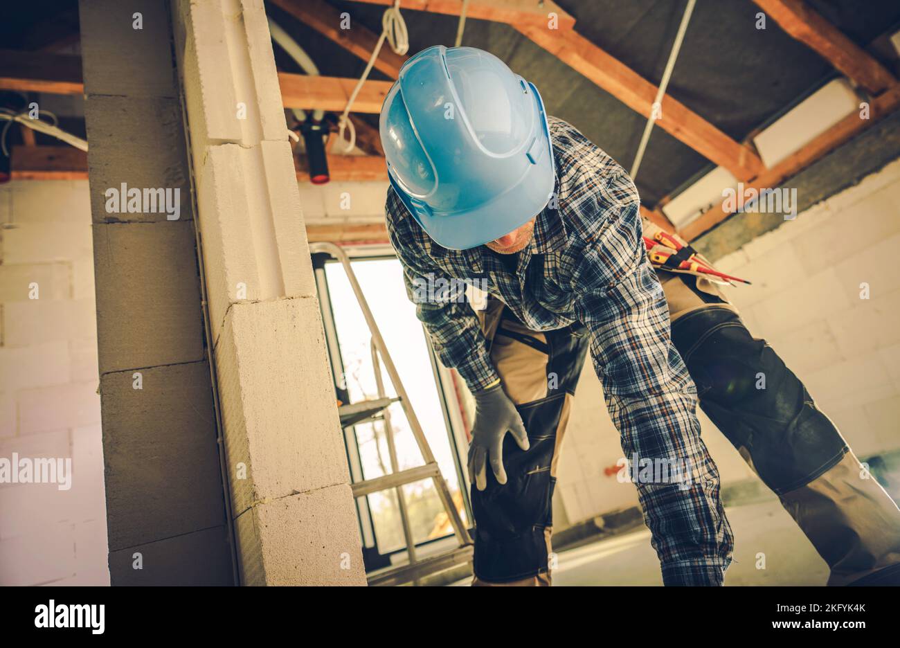 Lavoratore di costruzione professionale in Blue Hard Hat che si piega verso il basso per i suoi utensili. Casa residenziale edificio sito in background. Foto Stock