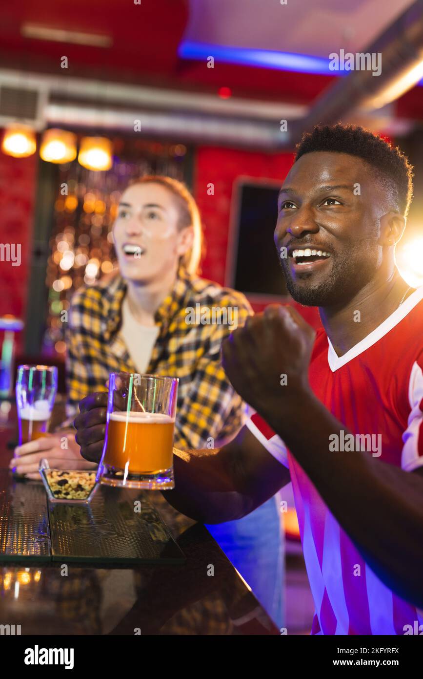 Verticale di amici maschili e femminili diversi ed eccitati che reagiscono al gioco di sport che si esibisca in un bar Foto Stock