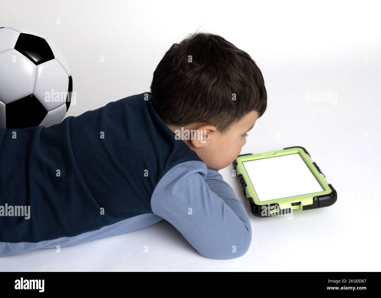 Ragazzo maschio di circa 4 anni e la sua palla di calcio sdraiata sul pavimento guardando il suo tablet. (Schermo vuoto) Foto Stock