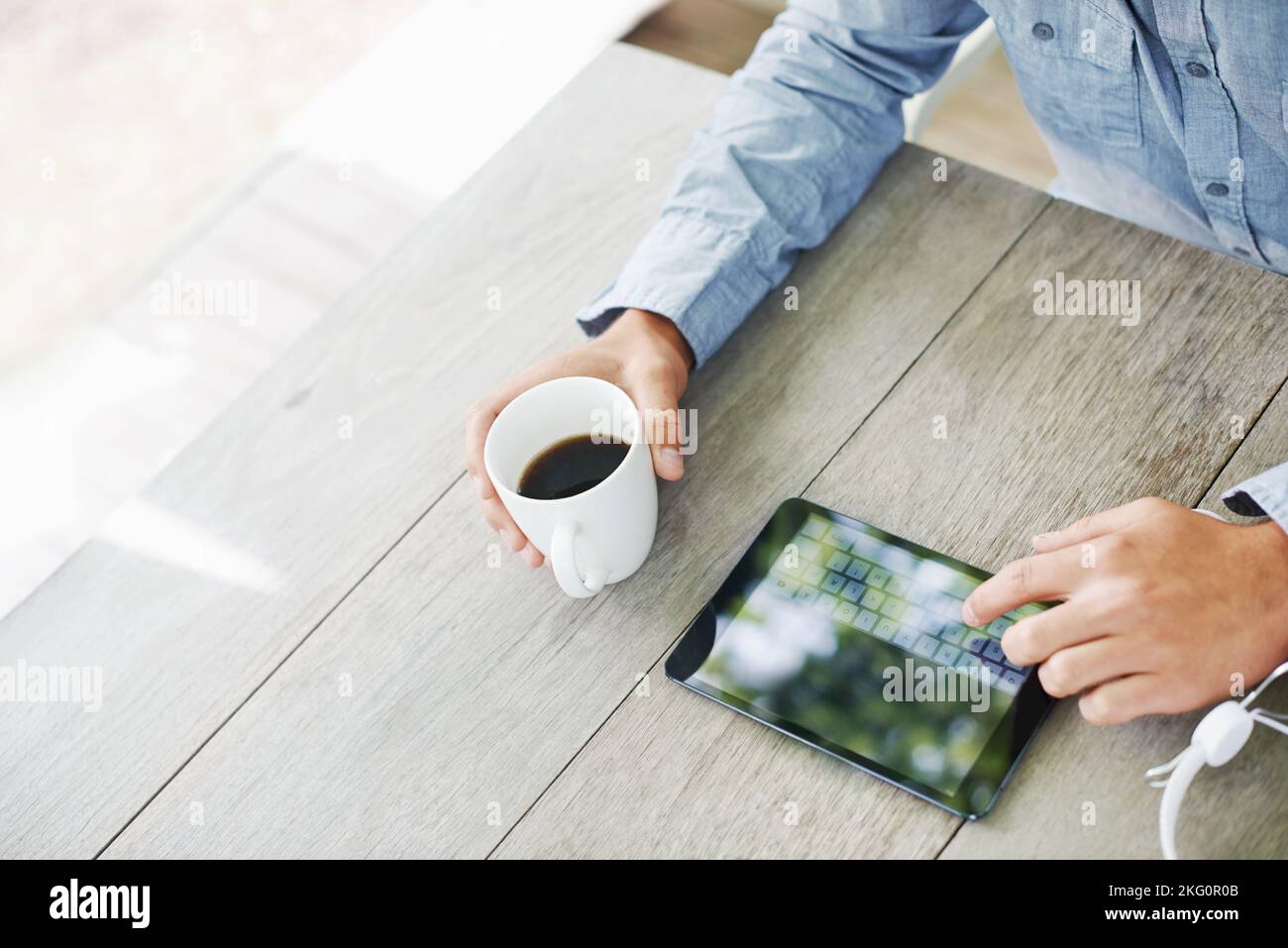 Inizia la tua giornata nel modo tecnologico: Un uomo che usa un tablet digitale mentre beve il caffè e le sue cuffie che gli sono accanto. Foto Stock