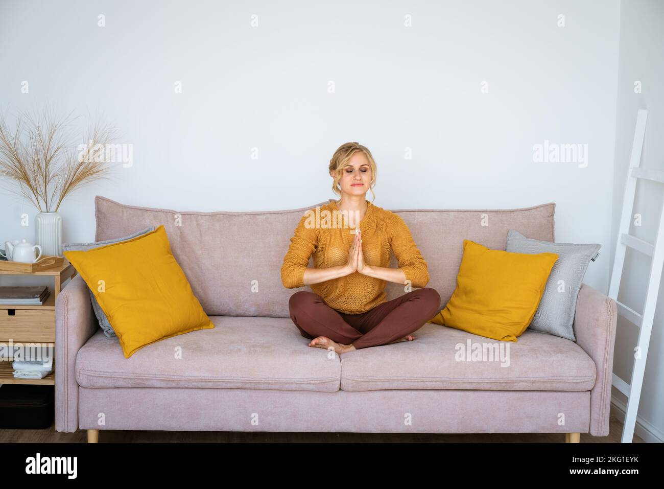 Giovane donna calma meditando in posizione loto facile a casa, seduta sul divano, tenendo le mani in un gesto di yoga, pratica yoga in un accogliente salotto, esercizio di respirazione aria, senza stress, crescita spirituale Foto Stock