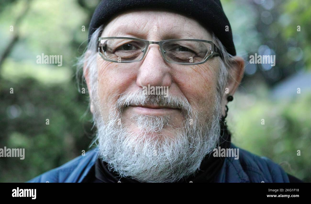 Ritratto di un uomo anziano di 68 anni che indossa abiti blu e un cappuccio nero. Ha una barba, occhiali e orecchie nere. Lo sfondo è sfocato Foto Stock