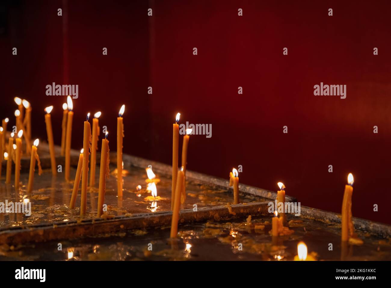 Candele di cera sottili illuminate in piedi nei portacandele sul tavolo della chiesa contro un muro rosso, concetto di religione Foto Stock