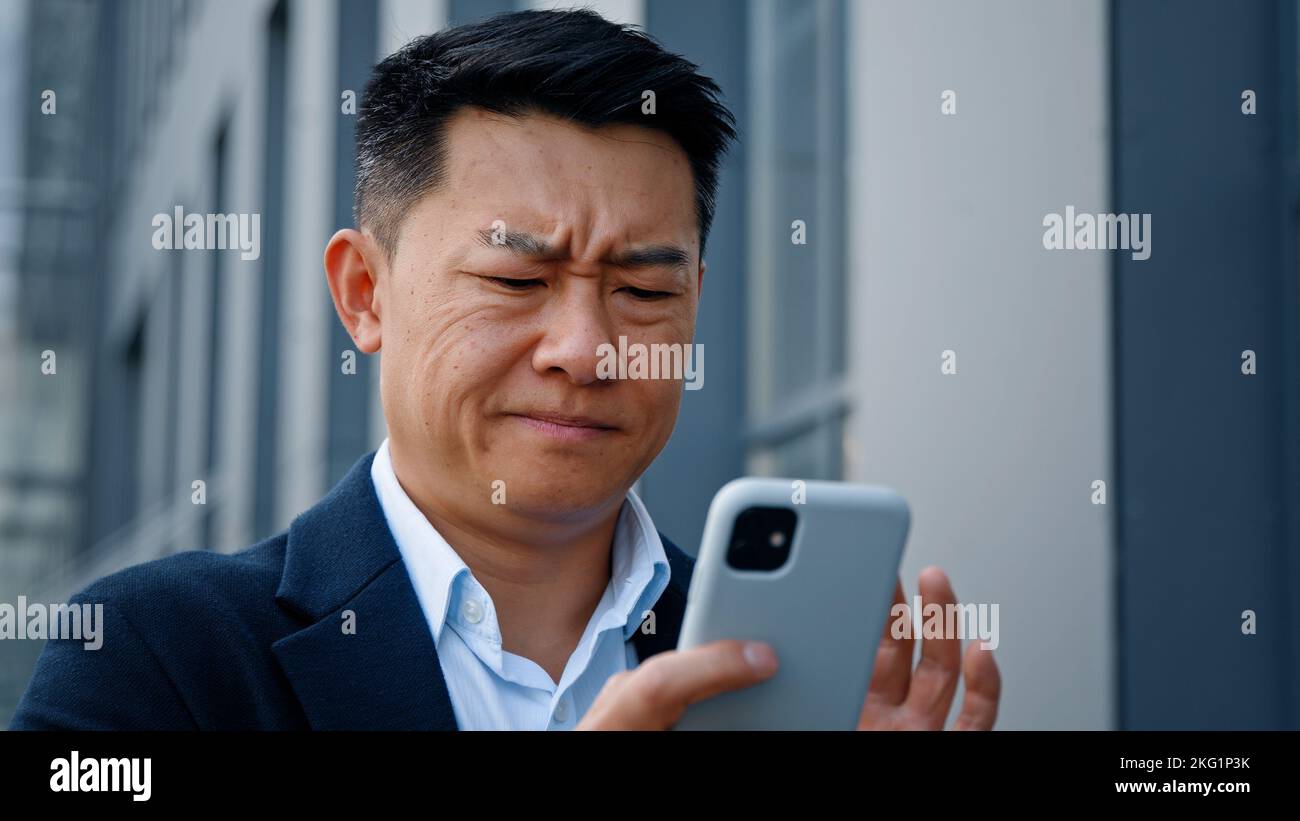 Primo piano arrabbiato preoccupato sottolineato asiatico di mezza età 40s uomo utente imprenditore datore di lavoro imprenditore uomo cercando di navigare sul telefono cellulare rotto all'aperto in città Foto Stock