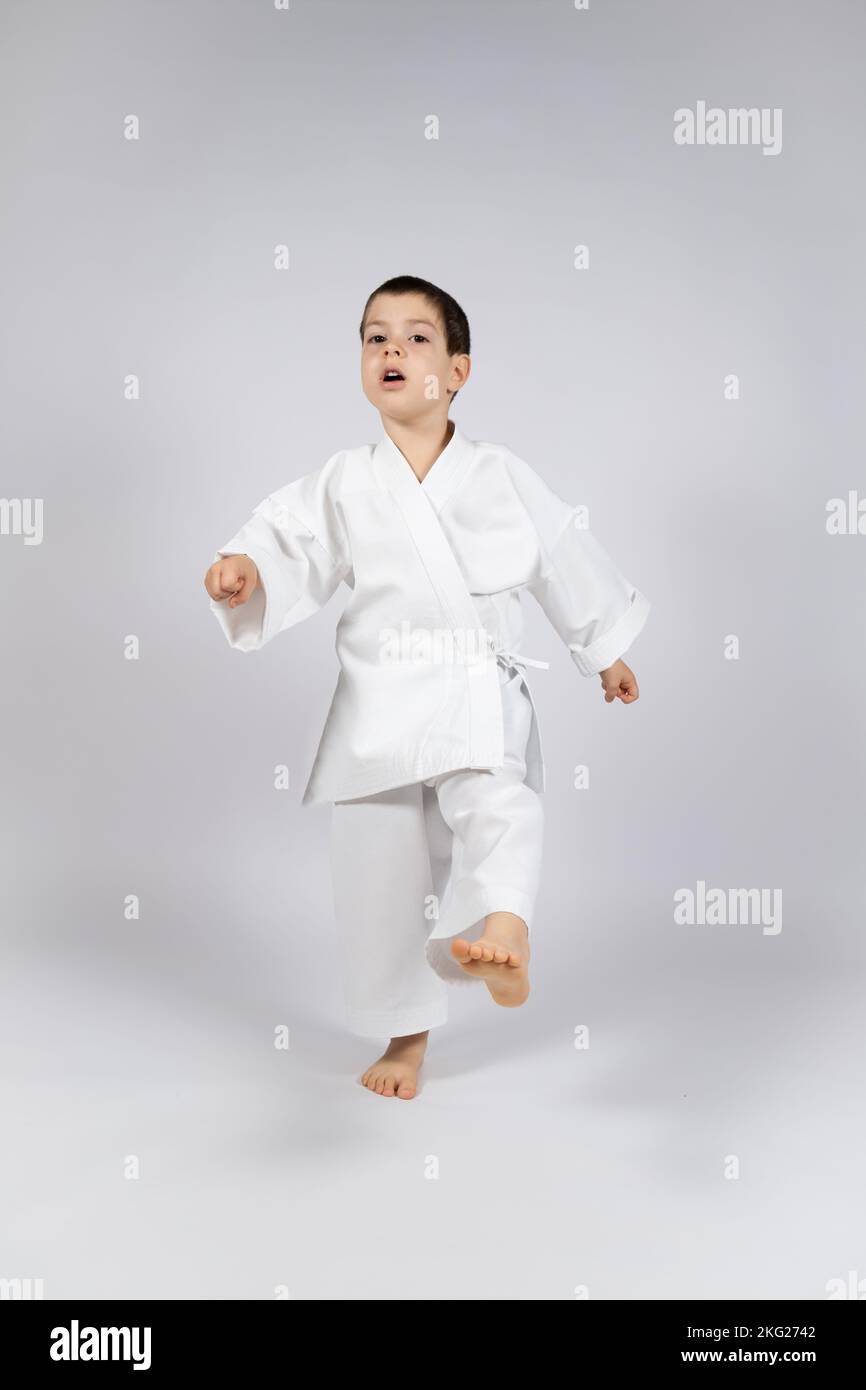 Un ragazzino in un kimono pratica il karate su uno sfondo bianco, calciando in avanti. Foto Stock