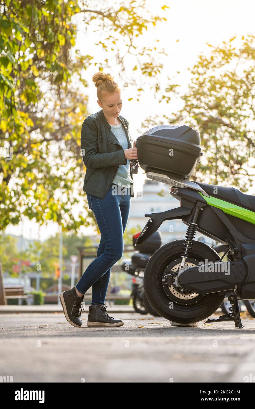 Donna dai capelli rossi che apre il tronco della sua moto parcheggiata Foto Stock