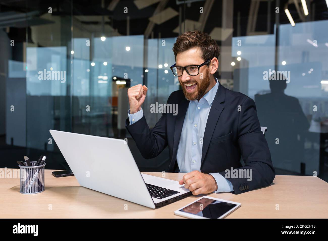 Uomo d'affari di successo che celebra la vittoria e il buon successo aziendale, il capo che utilizza il notebook per leggere la notifica dallo schermo e tenere la mano su un gesto trionfale sorridente. Foto Stock