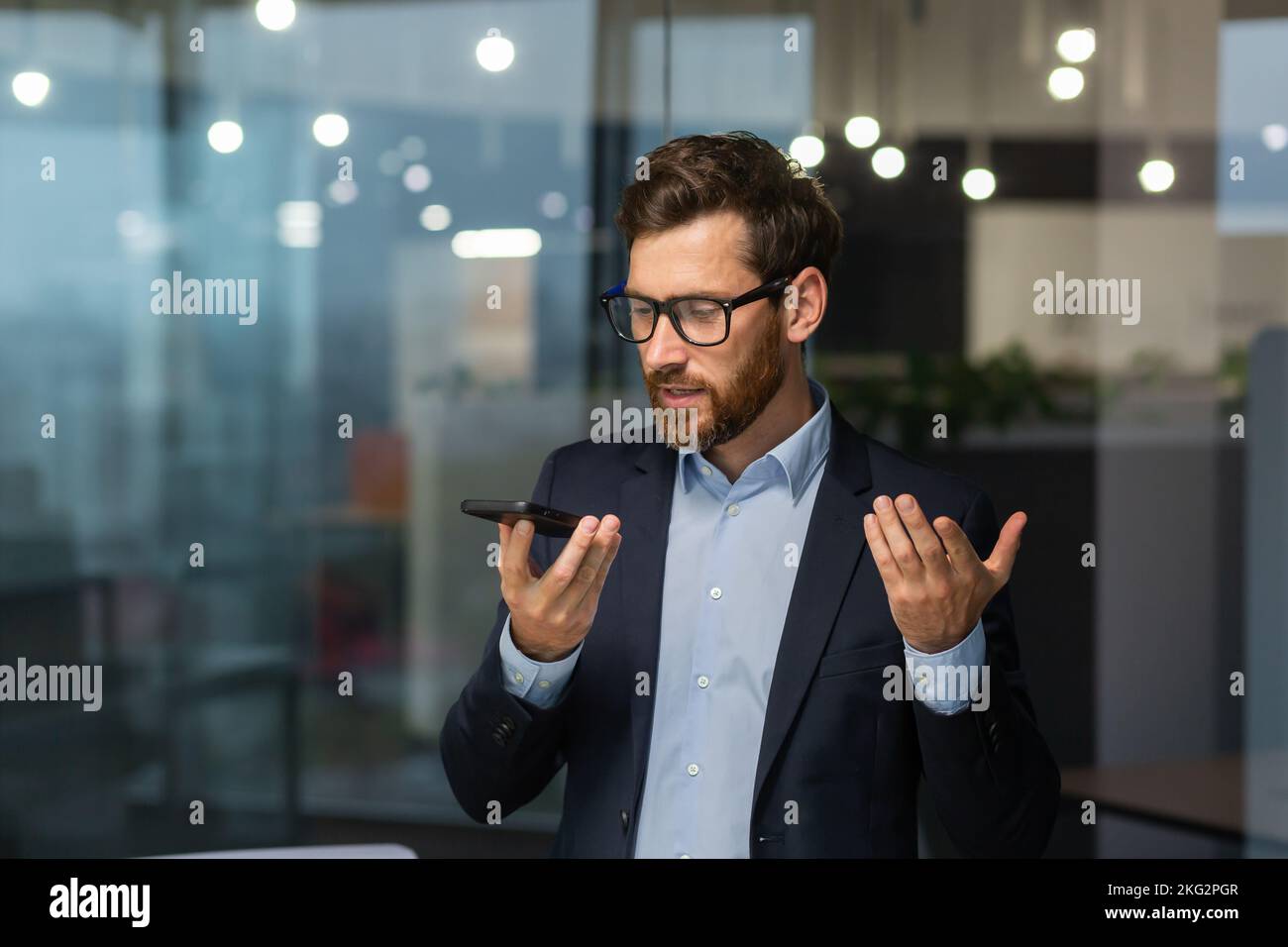 Un uomo d'affari amichevole e sorridente in ufficio che registra un messaggio audio, un uomo che utilizza l'app per smartphone per la comunicazione, un investitore in tuta da lavoro vicino alla finestra all'interno dell'edificio. Foto Stock