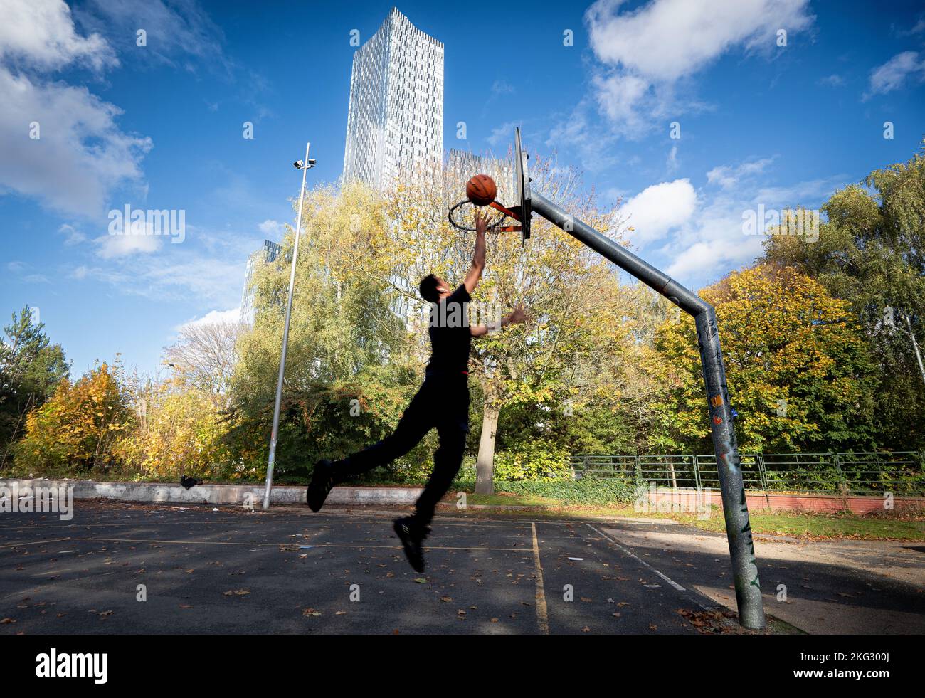 Il giovane uomo salta giocando a basket Hulme Park all'ombra della Torre Sud del nuovo sviluppo del centro città. Manchester UK Picture credit garyroberts/World Foto Stock