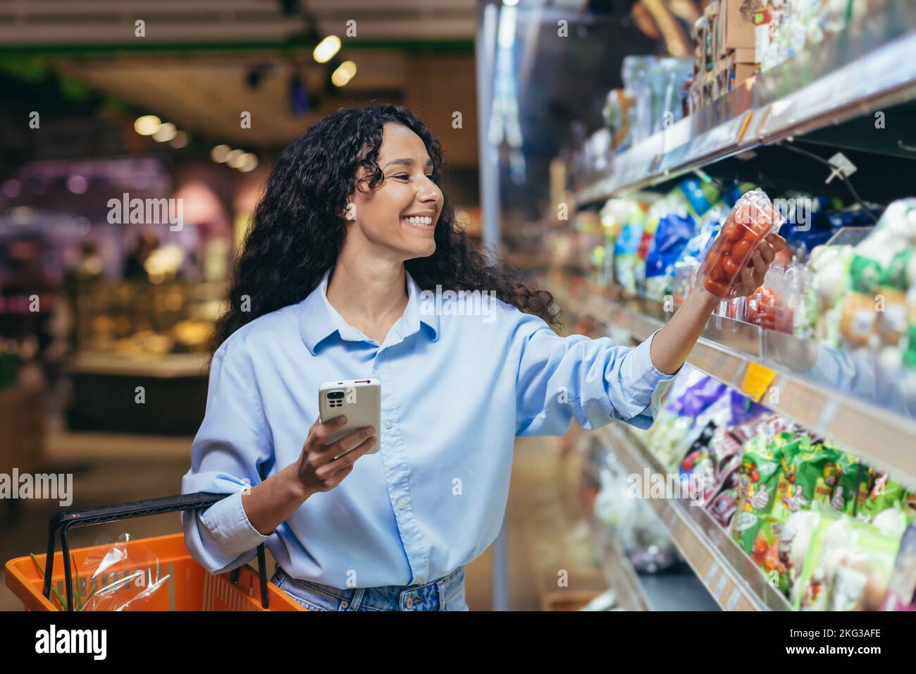 Donna acquirente felice con il telefono nel reparto della drogheria che naviga la lista della spesa in app online, scegliendo pomodori verdure nel reparto della drogheria. Foto Stock