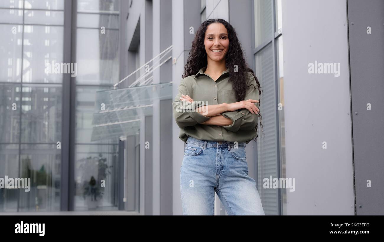 Nel centro città sfondo in piedi singolo sorridente felice fiero 30s donna in blusa casual forte posa fiduciosa con braccia incrociate sguardo femminile Foto Stock
