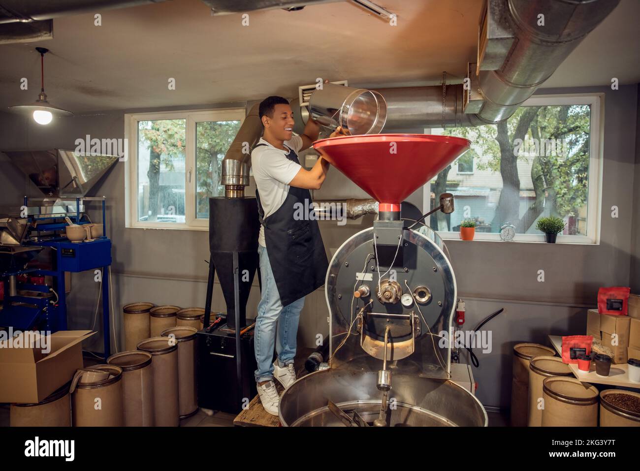 Un maestro di arrostimento allegro che aggiunge i chicchi di caffè nella macchina per la tostatura Foto Stock