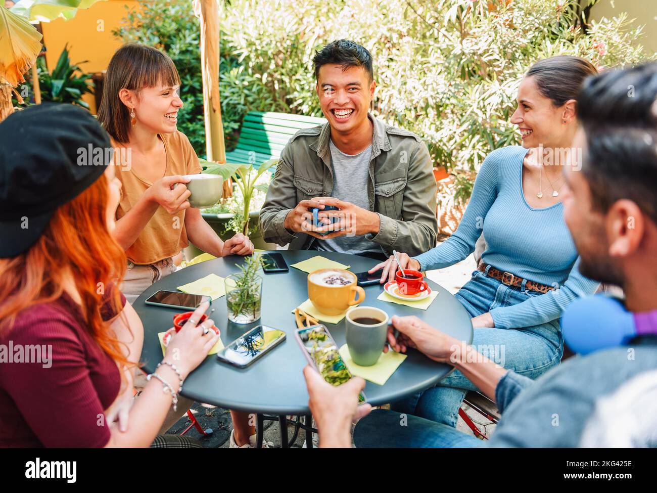 Da sopra gruppo di uomini e donne multirazziali allegri che ridono di scherzo mentre si siedono intorno al tavolo e bevono caffè fresco il giorno di fine settimana di estate dentro Foto Stock