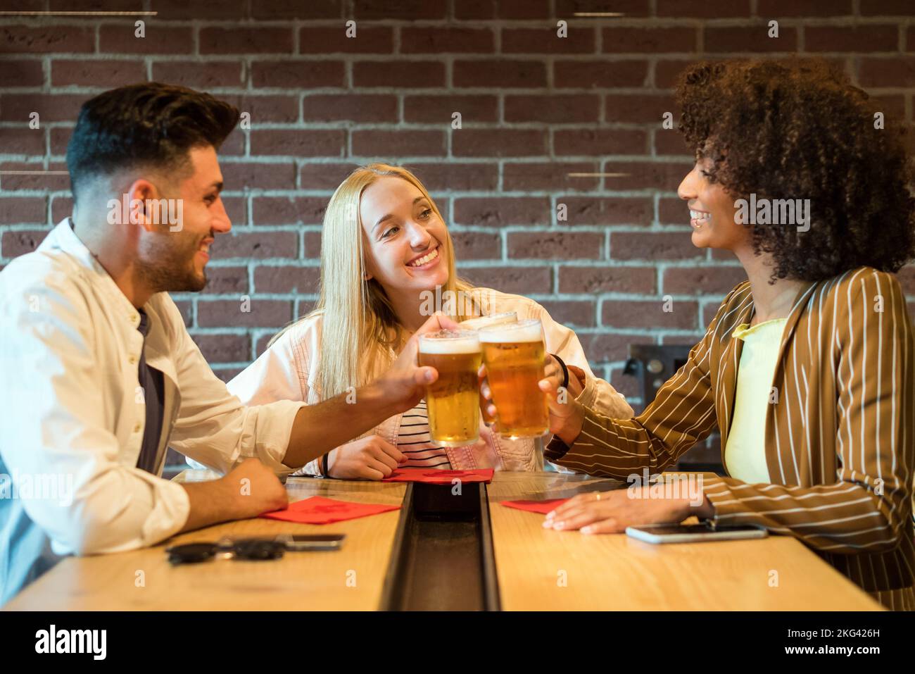 Sorridente allegria multirazziale con bicchieri di birra schiumosa fredda mentre si siede al tavolo nel pub vicino al muro di mattoni Foto Stock