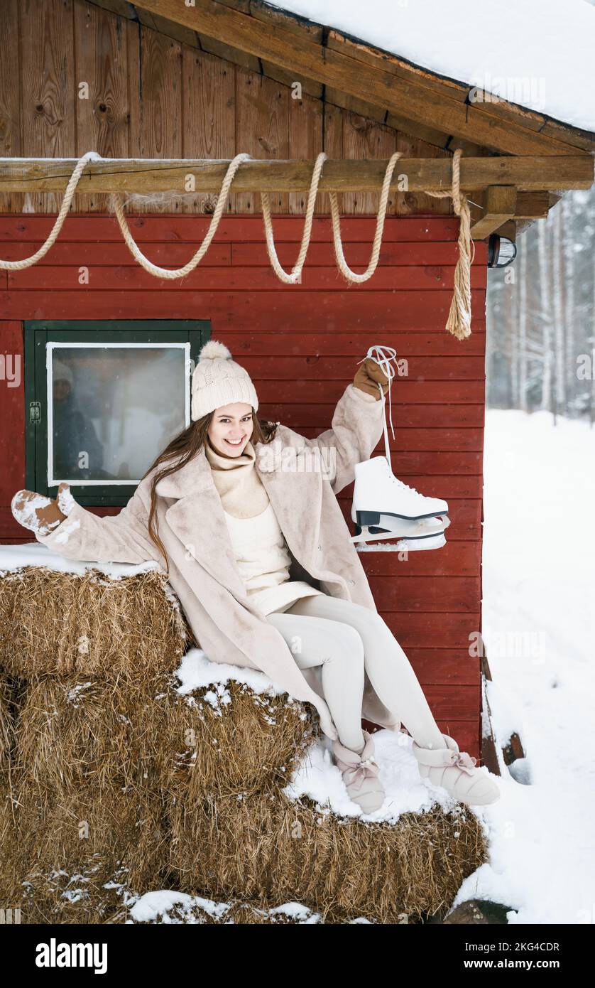 Giovane bella ragazza su balle di fieno vicino fienile con pattini. Inverno freddo innevato in campagna. Camminare, divertirsi, ridere in abiti eleganti, pelliccia co Foto Stock