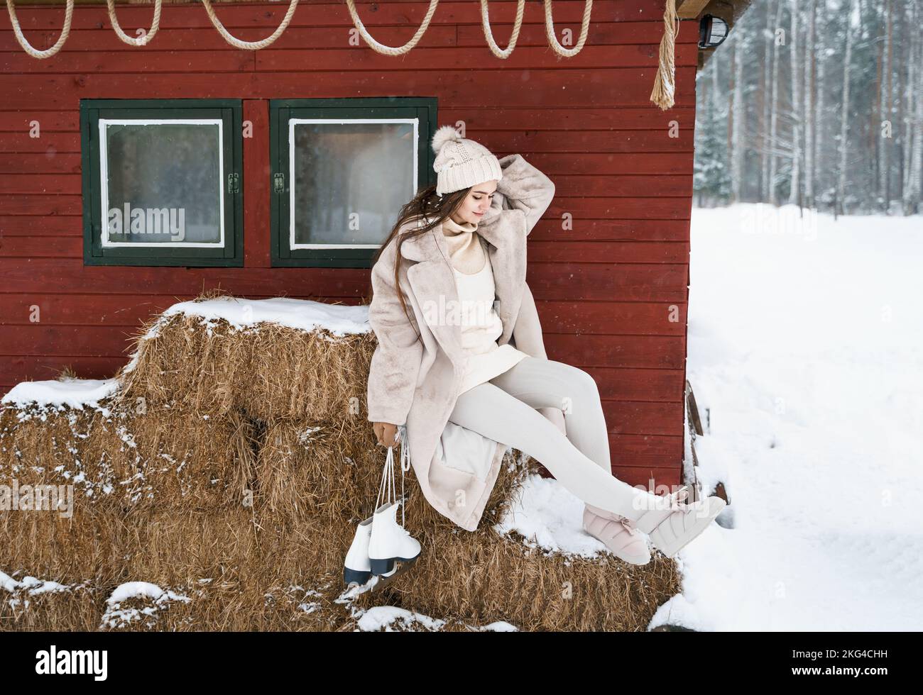 Giovane bella ragazza su balle di fieno vicino fienile con pattini. Inverno freddo innevato in campagna. Camminare, divertirsi, ridere in abiti eleganti, pelliccia co Foto Stock