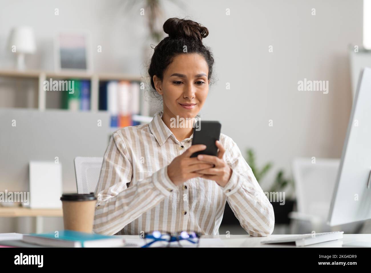 Sorridente giovane lavoratrice che usa lo smartphone e legge il messaggio, seduto al tavolo con il pc all'interno dell'ufficio Foto Stock