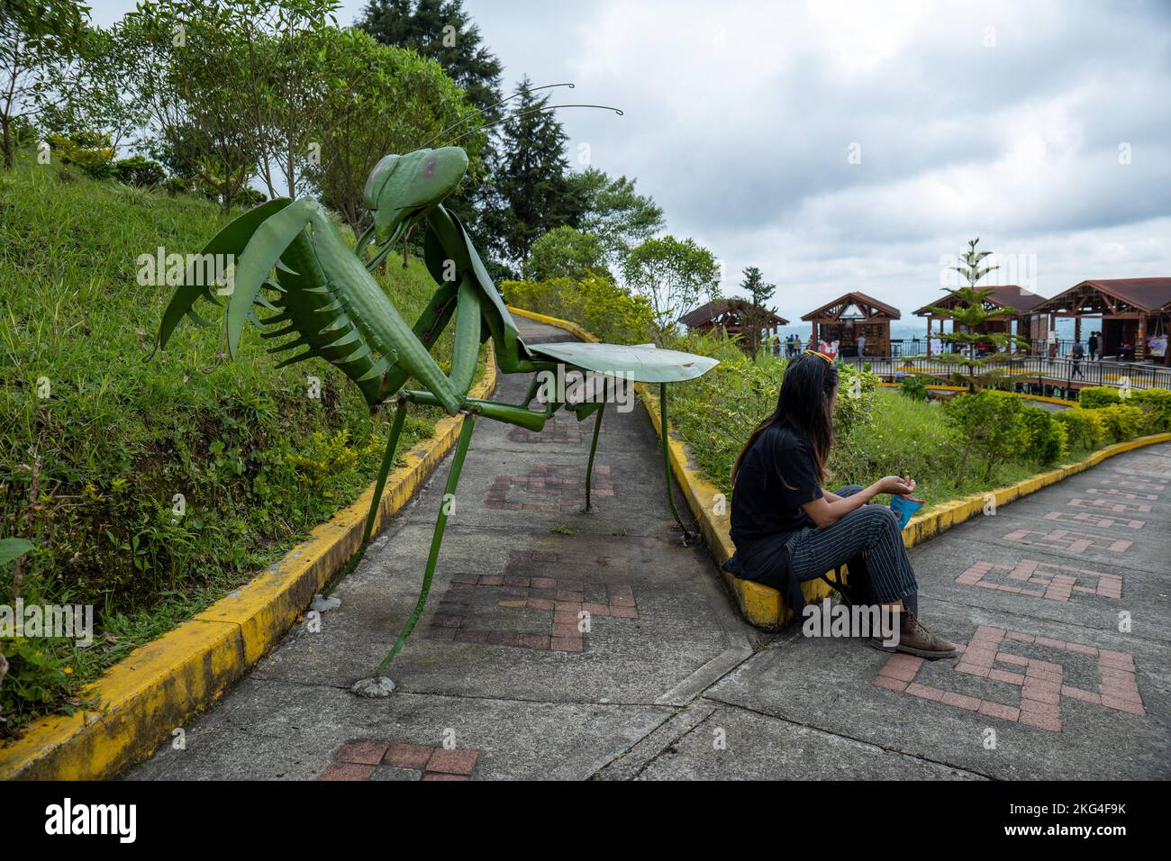 Filandia, Quindio, Colombia - Giugno 5 2022: Giovane Donna siede e spuntini di un pezzo artistico di metallo gigante Mantis contro un cielo grigio Foto Stock