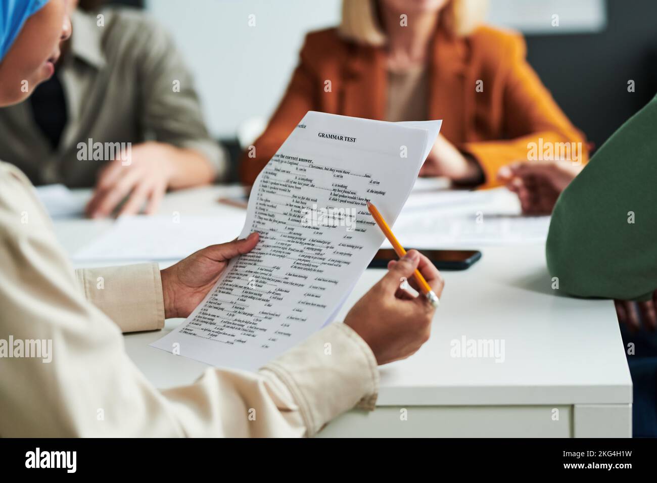 Focalizzazione selettiva sulle mani di una studentessa musulmana con matita che guarda attraverso compiti di test grammaticale contro insegnanti e giovani uomini Foto Stock