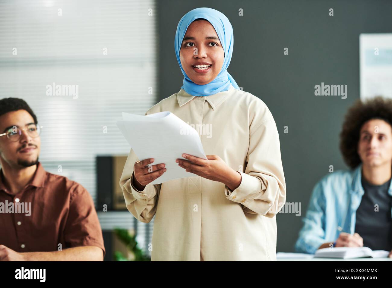 Giovane donna musulmana fiduciosa che parla in hijab blu facendo un discorso o una relazione al seminario mentre si è in piedi contro due uomini interculturali Foto Stock
