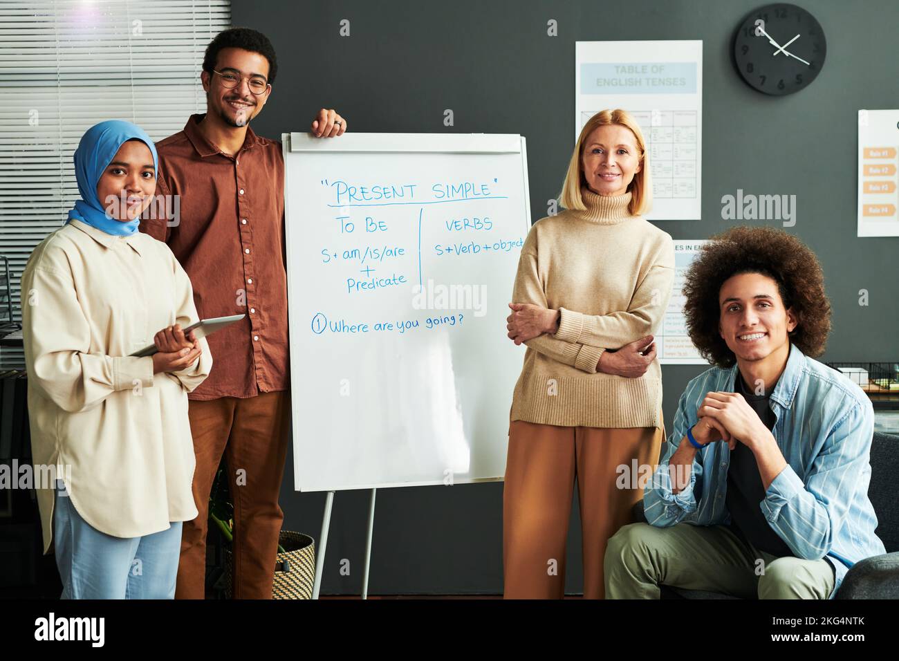 Diversi giovani studenti interculturali felici e biondo maturo insegnante che circonda lavagna bianca con spiegazione del presente semplice teso Foto Stock