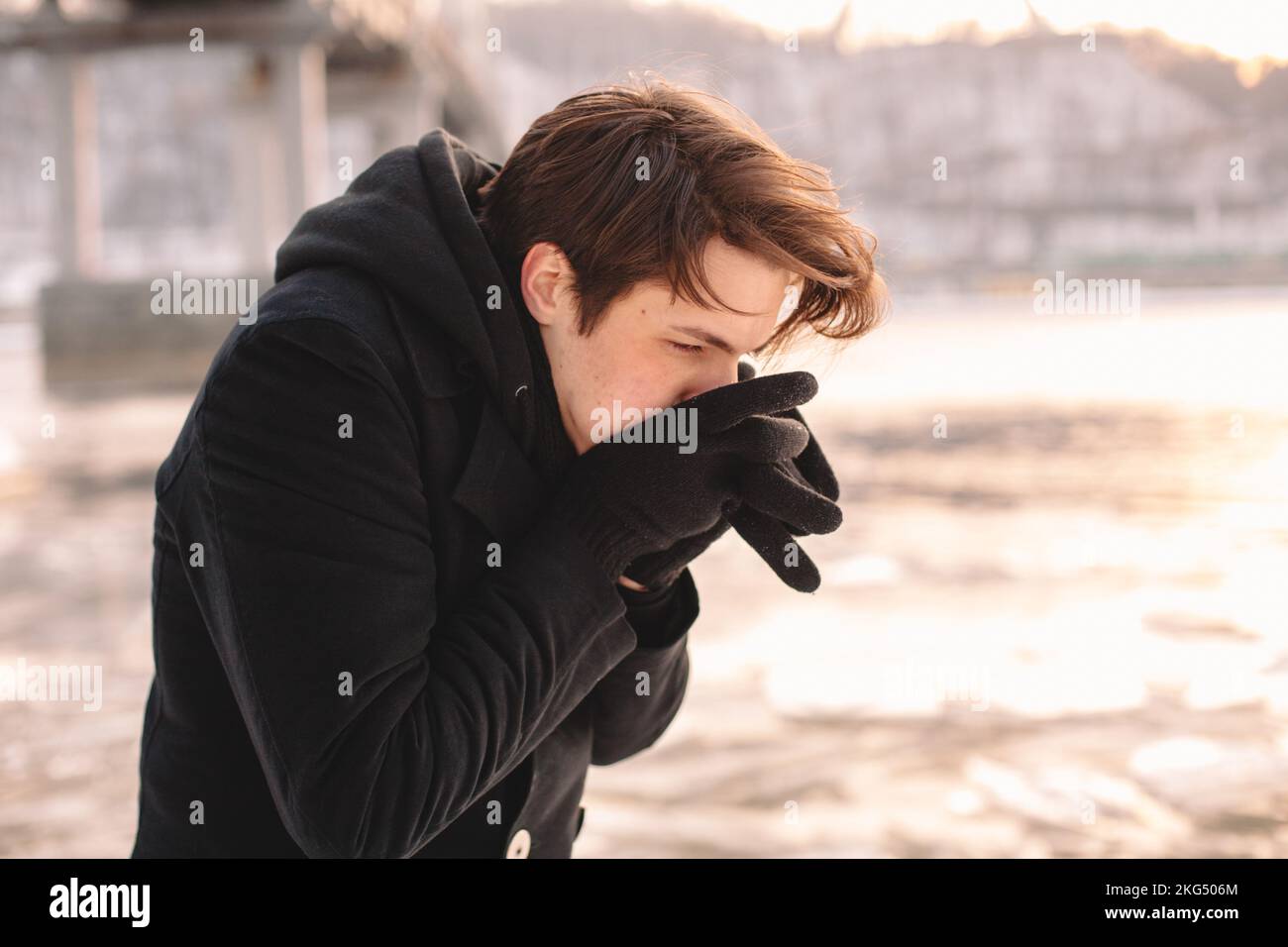 Giovane uomo che scalda le mani mentre si sente freddo in piedi dal fiume durante il tempo gelido in inverno Foto Stock