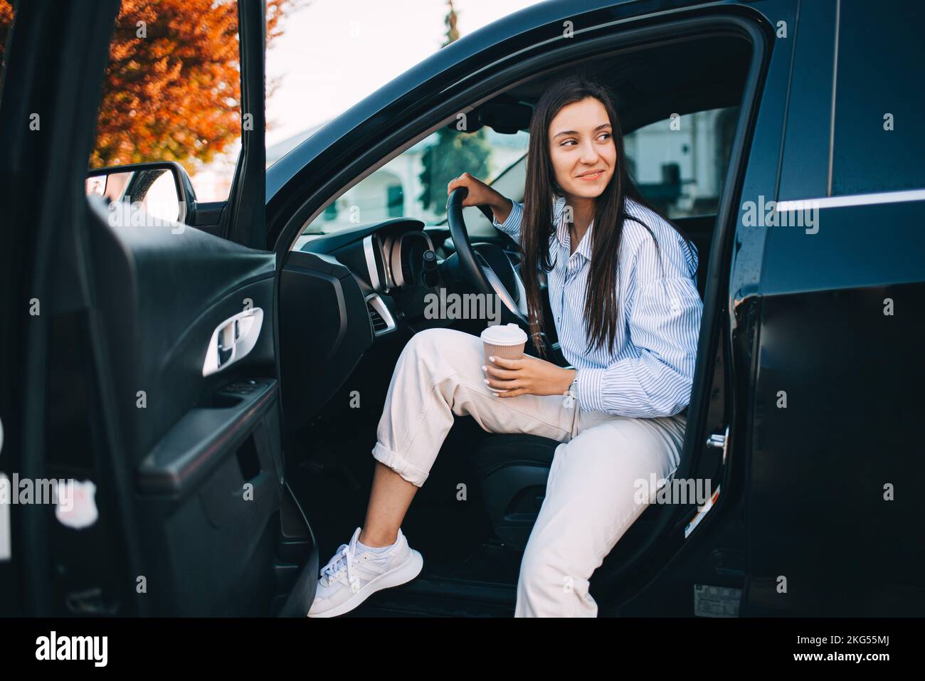 Una donna felice e attraente che tiene una tazza di caffè e che esce dalla sua auto moderna Foto Stock