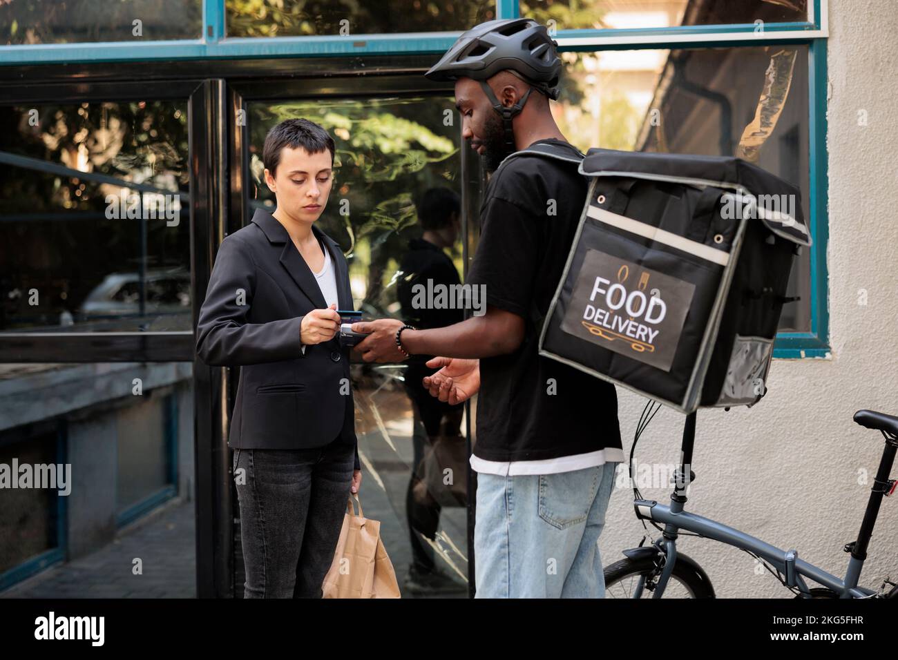 Servizio di consegna cibo in ufficio, pagamento tramite carta di credito presso il terminal POS. Corriere afro-americano in bicicletta che consegna il pasto da asporto del ristorante, dipendente dell'azienda che paga senza contatto per il pranzo Foto Stock