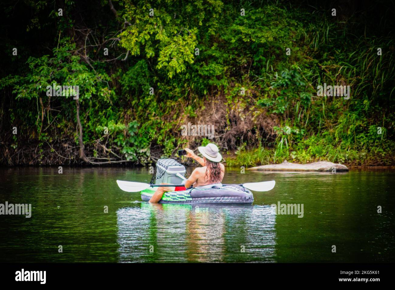 Una persona che galleggia lungo il fiume su una zattera di salvataggio con petto di ghiaccio e cappello bianco e remi che si videofanno con il telefono - riflessi e densa sfocatura Foto Stock