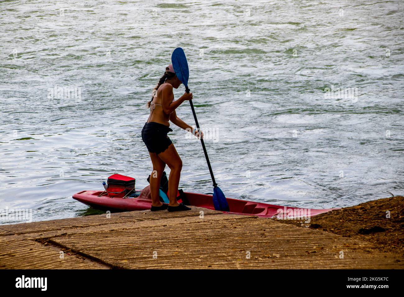 Donna adombrata in pantaloncini corti e berretto da bikini tira fuori i remi dal kayak tirati fino alla rampa di barca con l'acqua sullo sfondo Foto Stock