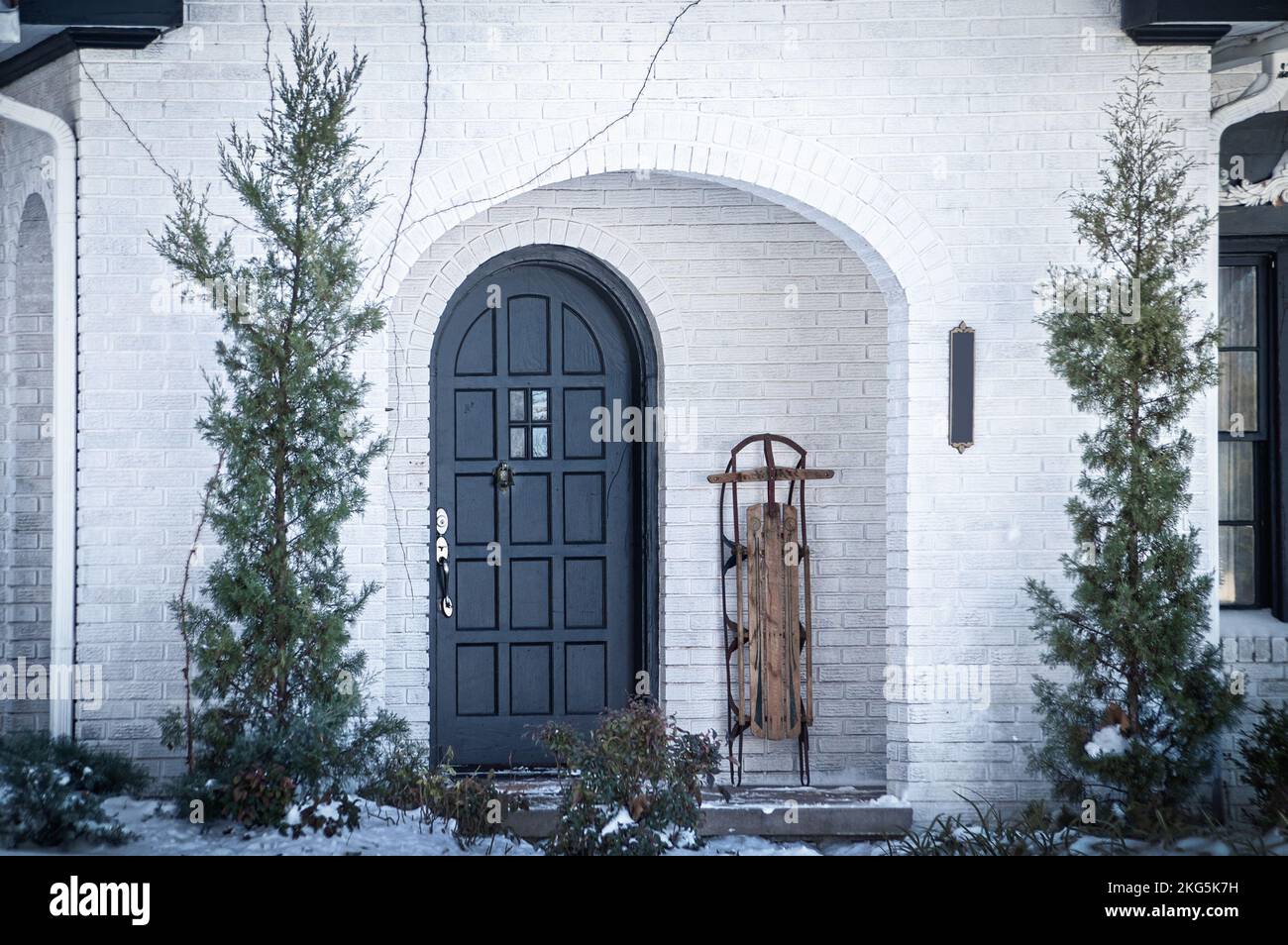 Giorno della neve - portico di casa in mattoni dipinti di bianco con porta anteriore ad arco e slitta punteggiata nelle vicinanze - alberi Evergreen a ogni lato Foto Stock