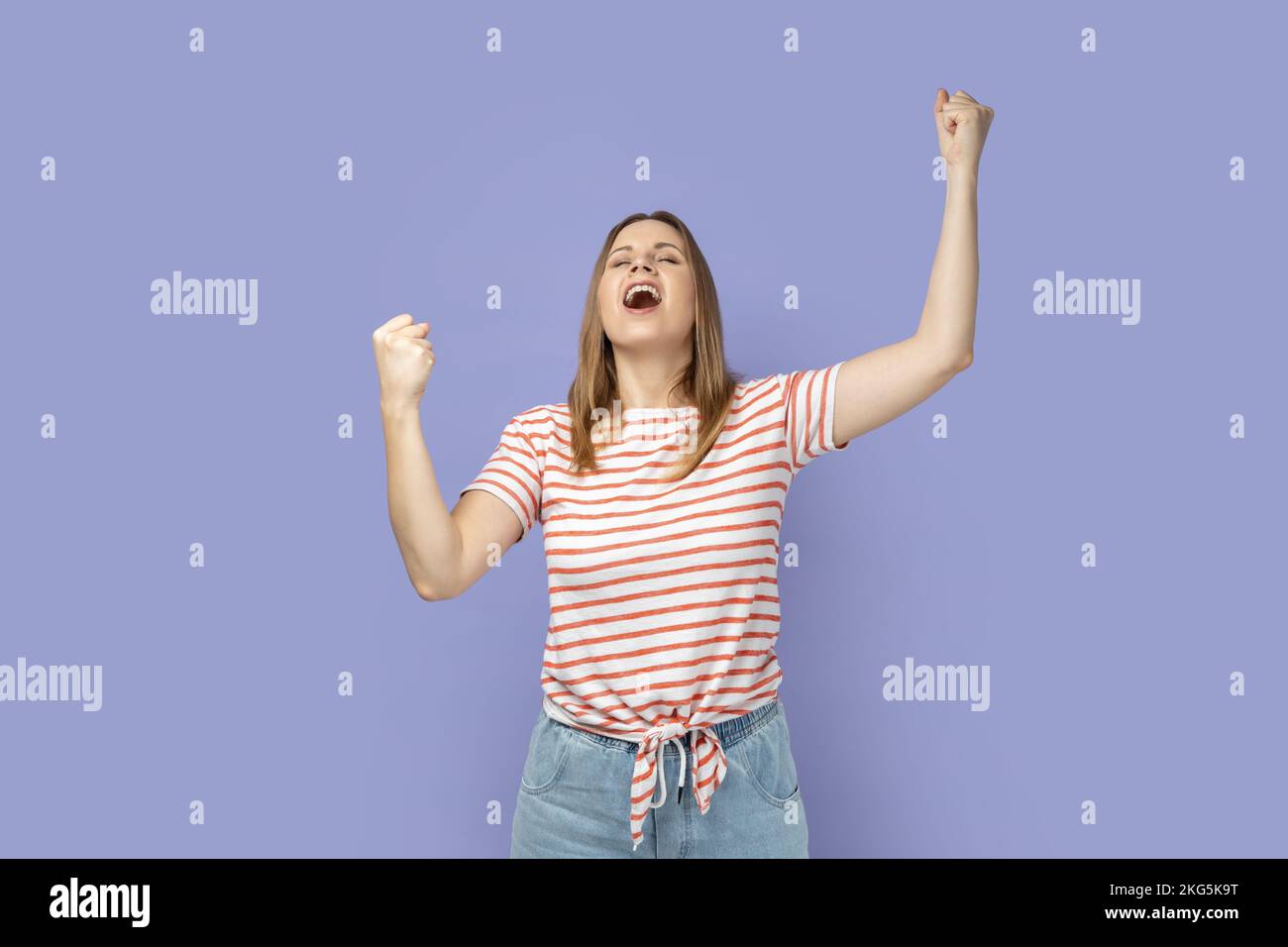 Ritratto della donna bionda che indossa una T-shirt a righe che mostra un gesto di sì e urla celebrando la sua vittoria, il successo, i sogni si avverano, euforia. Studio al coperto isolato su sfondo viola. Foto Stock